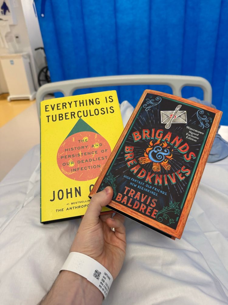 Holding two books in a hospital bed, one is ‘Everything is Tuberculosis’ by John Green, the second is ‘Brigands & Breadknives’ by Travis Baldree.