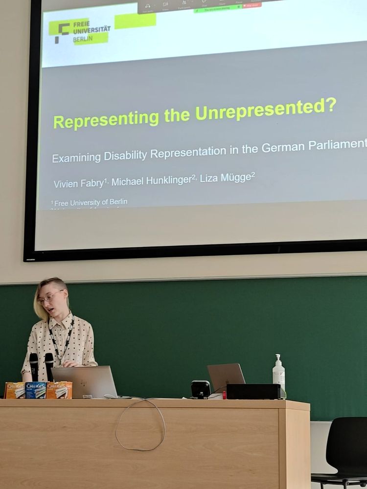 Picture of a person with glasses and blonde hair, wearing a shirt, standing in front of a projector screen. The screen shows a presentation title slide saying "Representing the Unrepresented? Examining Disability Representation in the German Parliament"