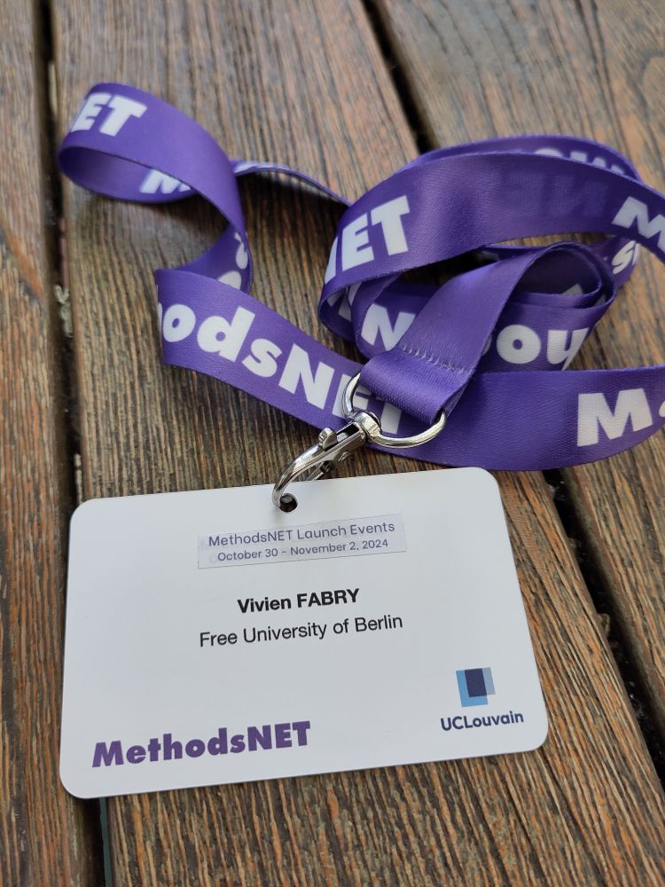 A conference badge from the MethodsNet conference. It has a purbple lanyard and lays on a wooden table.
