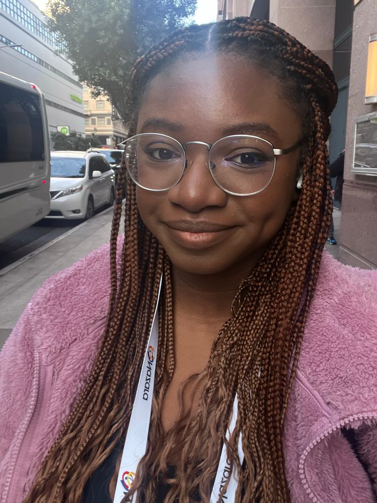 Myself smiling outside in sunny weather. I’m wearing my GDC 2025 badge and a pink sweater. 