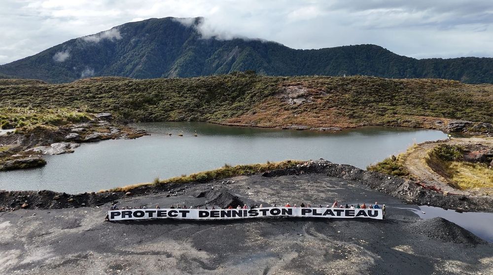 Wide shot of banner reading protect denniston plateau, with a lake in background