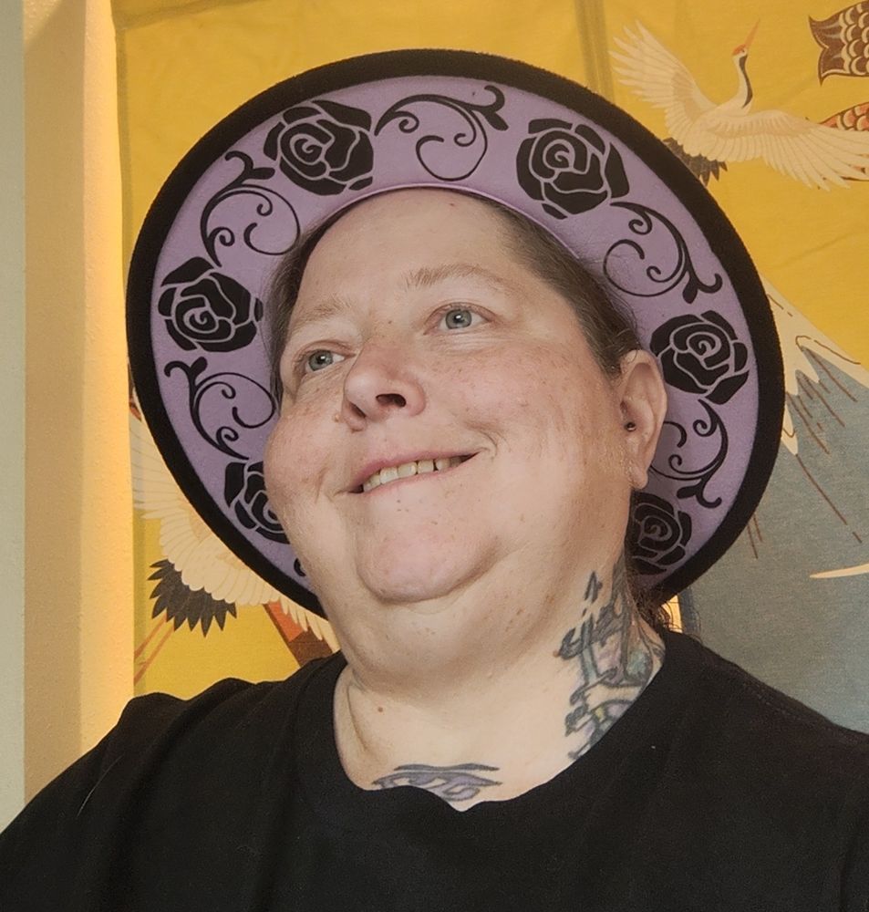 Lanta in a black shirt and a wide brimmed black hat with lavender patterned with black roses underneath the brim.