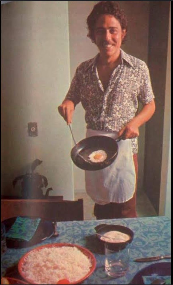 chico buarque sorrindo segurando uma frigideira com um ovo frito diante duma mesa posta 