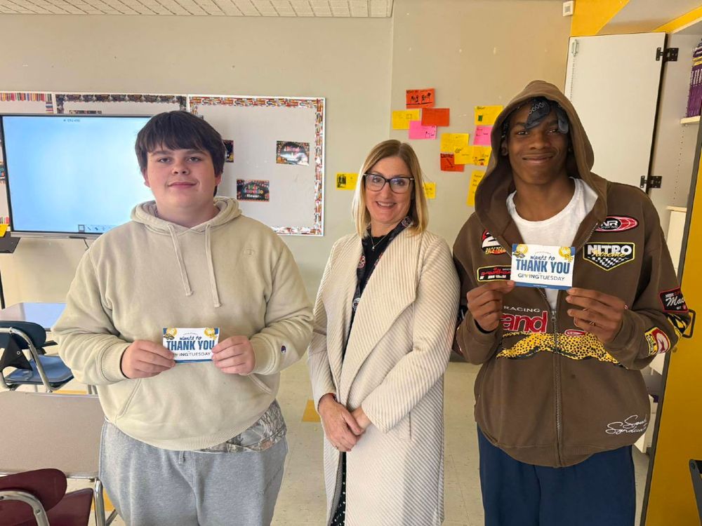 Two students holding "Thank You" certificates stand beside a smiling teacher in a classroom with colorful posters and a whiteboard.