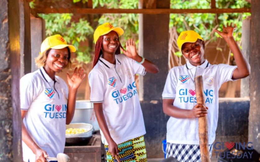Three smiling women in matching "Giving Tuesday" shirts and yellow caps wave while cooking. "Giving Tuesday" logo is visible.