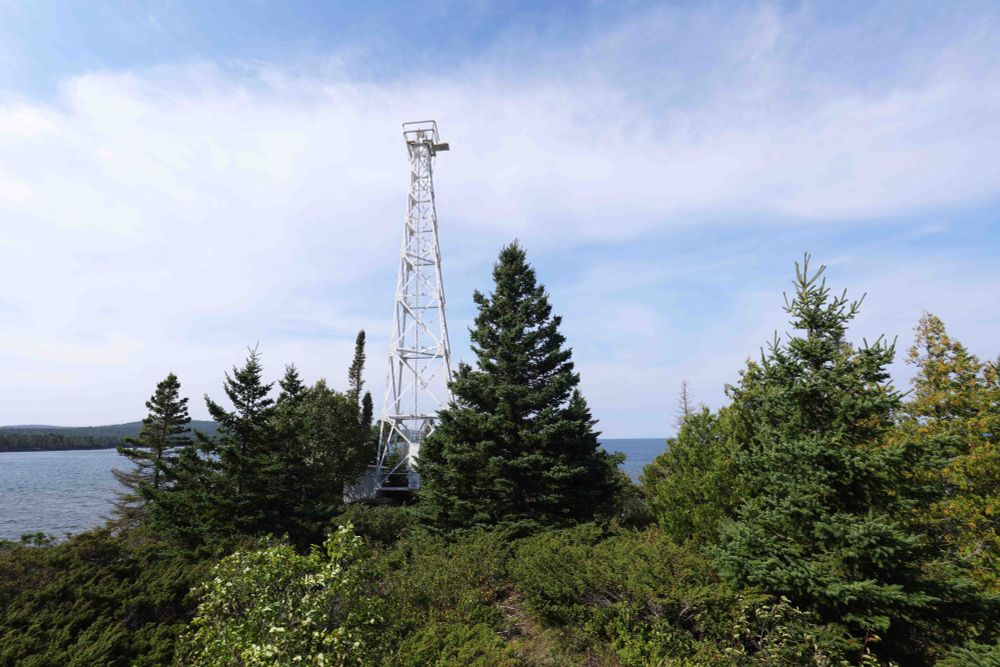 Actual lighthouse light on the shores of Lake Superior, a maybe 35' tall structure with a green light to ward ships away from the very rocky shore. It is surrounded by evergreens. 