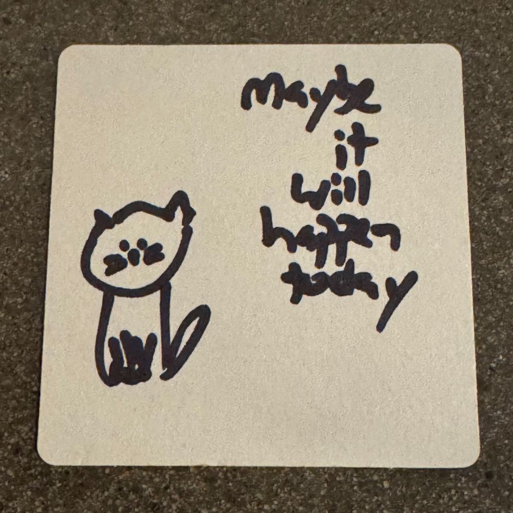 The blank back of a beer mat with a little sharpie drawing of a kitty cat on it and the words ‘maybe it will happen today’