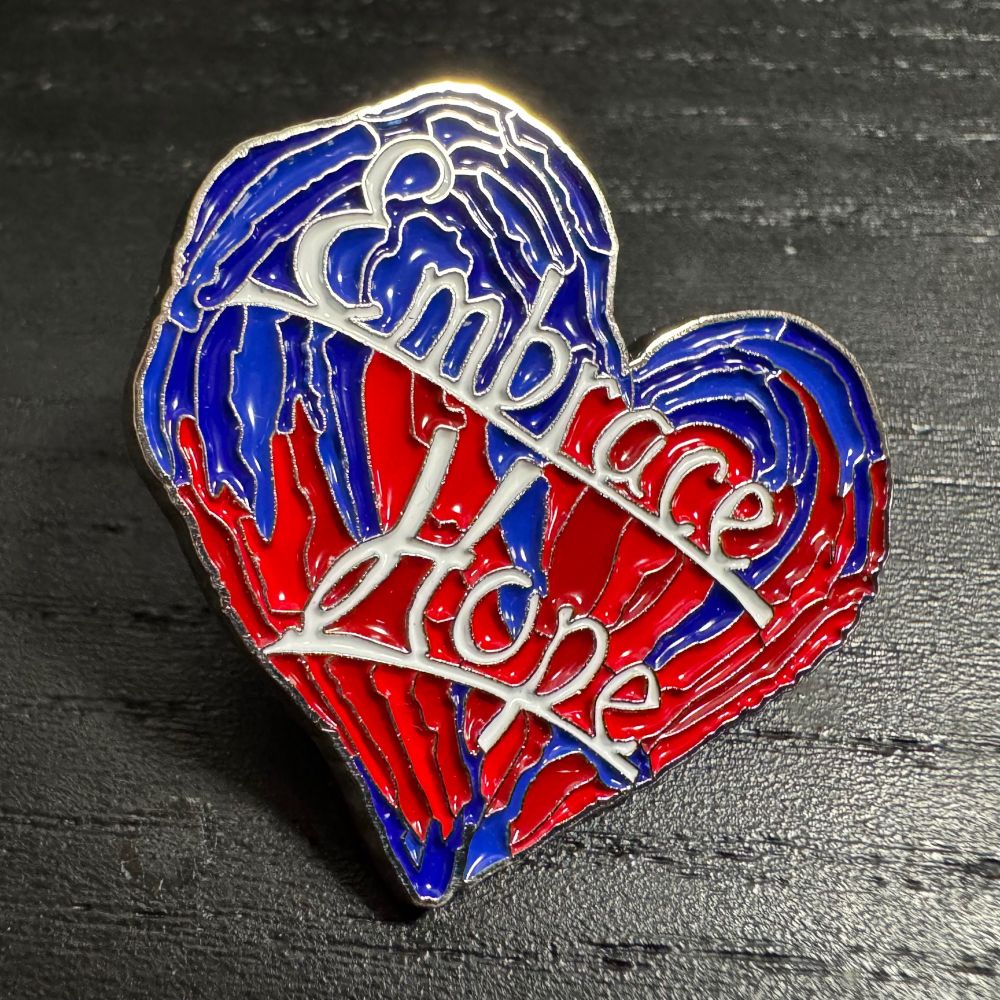An enamel pin in a blue and red heart design that has script lettering over top of it reading 'Embrace Hope'