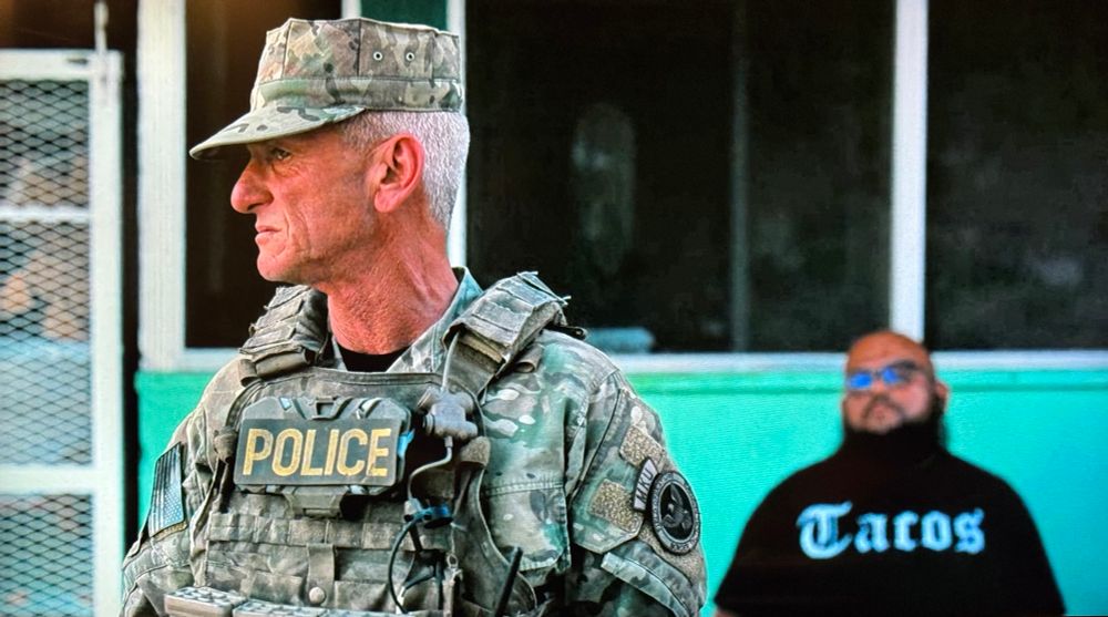 Screenshot from One Battle After Another. Sean Penn in the foreground wearing full camo BDUs with a POLICE badge across his chest, looking angry. Behind him, seated and a bit out of focus, a bearded Mexican gentleman wearing a black t-shirt with the word ’Tacos’ printed on it in white cholo gothic type. 
