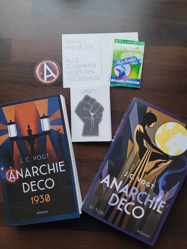 Paket der Vögte "unboxed". Signierte Exemplare von Anarchie Deco und Anarchie Deco 1930, "Flugschriften der Schwarzen Scharen", eine Postkarte (Vorderseite Anarchie Deco 1930 Cover, Rückseite " Damals wie heute gemeinsam gegen den Faschismus", Aufkleber mit dem Buchstaben A und eine Tüte Ahoi Brausepulver Waldmeister