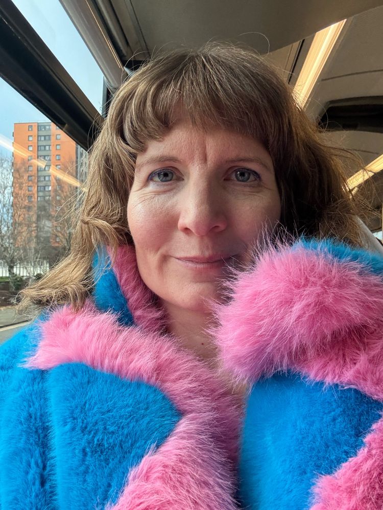 A selfie of me on a bus. I have brown/reddish wavy hair with a short fringe, and blue eyes. My coat is big, furry (fake fur of course!) and light blue with a baby pink trim. 