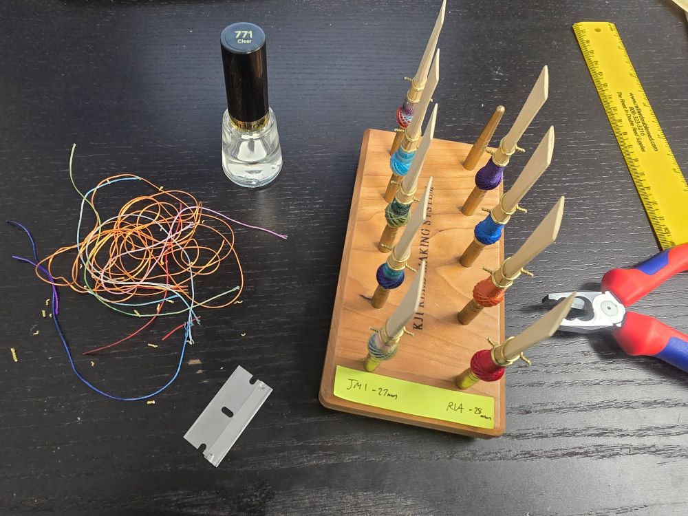 9 bassoon reed blanks of varying color wraps drying on a rack. Thread and wire clippings can be seen in a pile to the left, clear nail polish to the top left, a ruler and pliers to the right, and a razor blade at the bottom left.