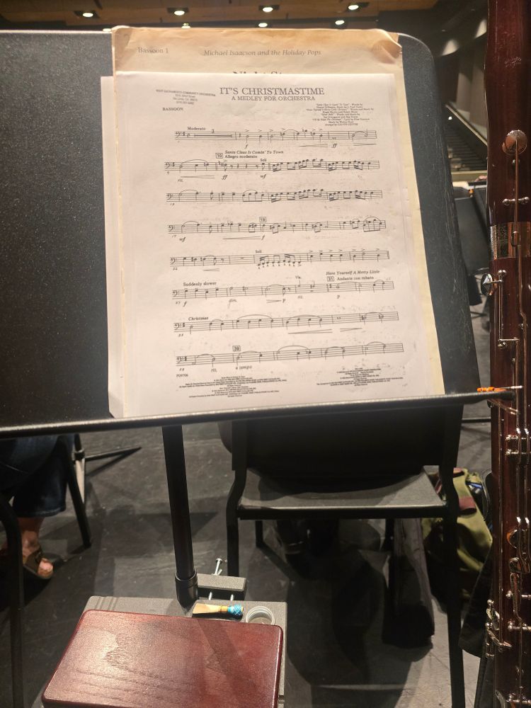 A first-person POV looking at a music stand with various pieces of sheet music on it. To the right side is a bassoon on an instrument stand.