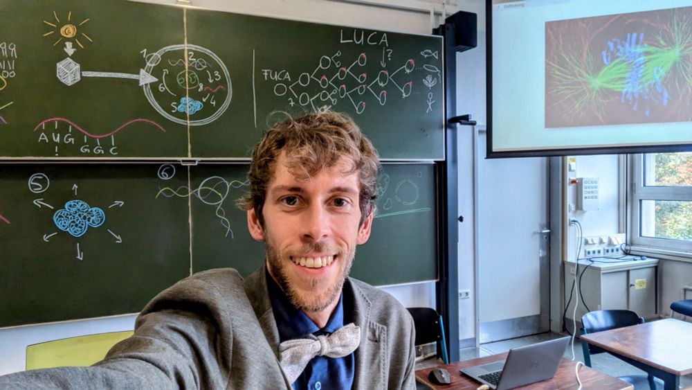 A guy with a bowtie in front of a black board. He looks happy after giving a biology lecture.