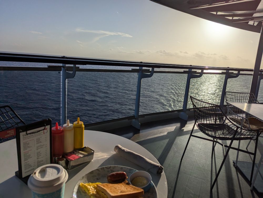 An ocean view from the back of a cruise ship as the sun rises. A table with coffee and a breakfast meal. 