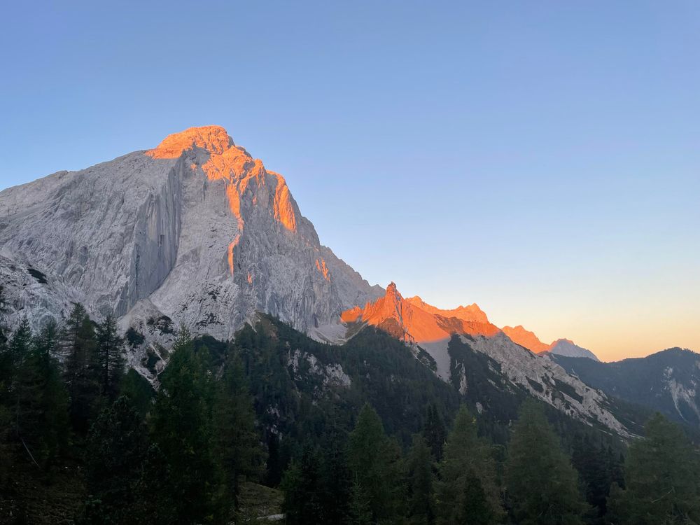 Grey limestone rock summits with orange/golden tips from sun beams above dark spruce forests. Clear blue sky