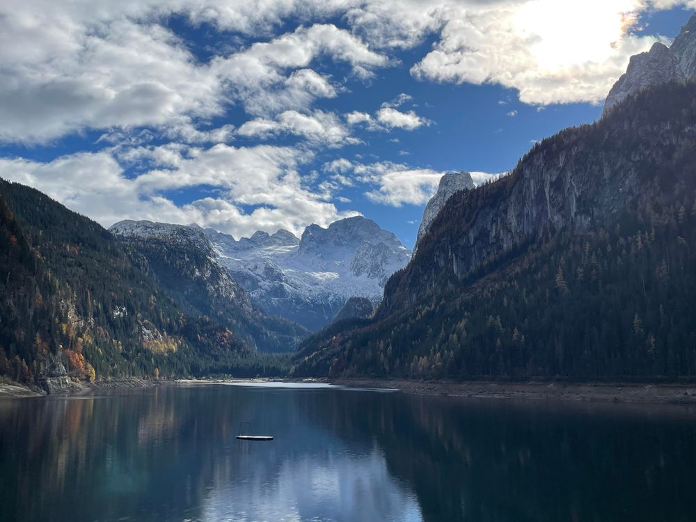 A snow covered, rugged Mountain range with a glacier in the middle ground that mirrors in a lake in the foreground. To the right are spruce forests in the dark shoddow with some limestone summits above, to the left spruce and some colourful deciduous trees lit by the sun. Blue sky with white clouds. The sun shines through a cloud in the upper right corner.