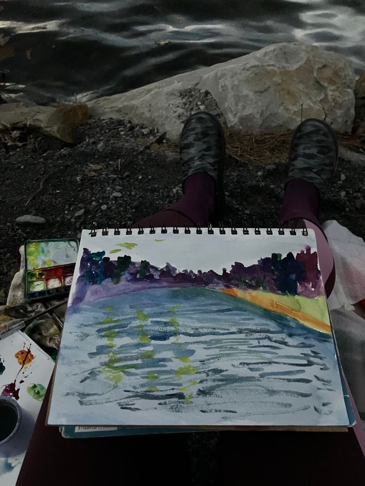 A picture showing my painting palette, a recycled yogurt jar of lake water for rinsing my brushes holding down my palette paper where I mix my colors, the painting notebook on my lap, warmer socks under my black and white camouflage shoes, the rocks and the lake at Keystone Lake State Park in southwestern Pennsylvania, about 1 hour from Pittsburgh. 