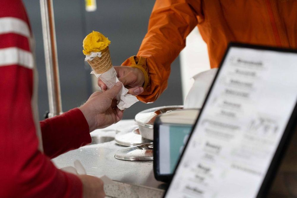 A close-up of an ice cream cone being handed to a person. The cone has two scoops: one bright yellow and one white with dark specks. The server in an orange jacket passes it across the ice cream cart counter, which has a menu and ice cream containers visible.