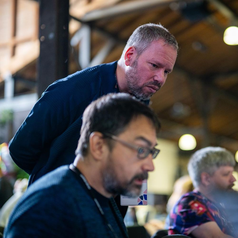 One person leans in to closely observe another working on a laptop at an indoor event. The background shows other attendees and a high wooden ceiling with exposed beams.