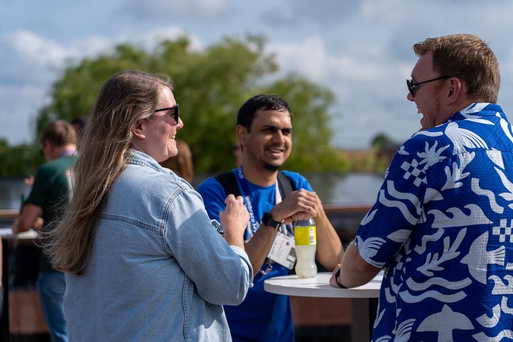 Three people chatting and smiling outdoors at a high table during a sunny event. They are wearing event lanyards and sunglasses, with trees and a lake in the background.