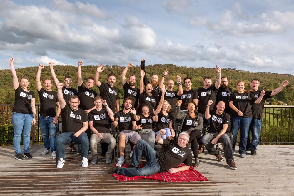 A large group of enthusiastic people wearing matching “CodeCabin 2018” black t-shirts pose outdoors on a wooden deck. Many have their arms raised in celebration. The group is surrounded by lush green hills under a partly cloudy sky, with one person playfully lying on a red picnic blanket in front.