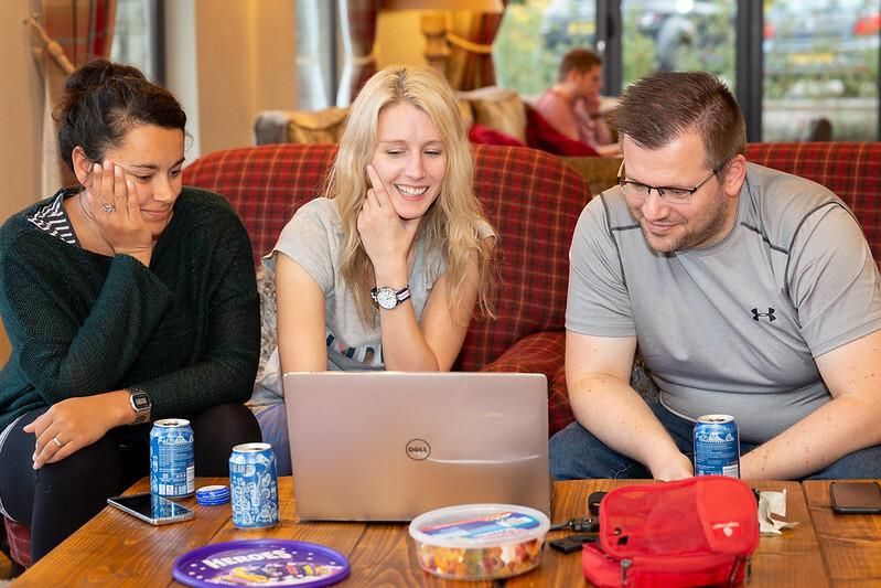 Three people sit on a cozy red plaid couch, smiling and focused on a Dell laptop. Two women and one man lean in, enjoying a relaxed collaboration. The table in front of them holds soda cans, snacks, and a red pouch. A casual, homey atmosphere is visible, with others chatting in the background.