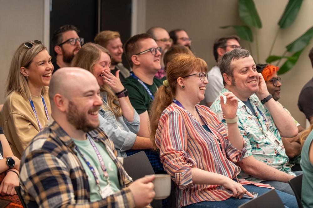 Audience seated indoors, laughing and smiling during a presentation at an Umbraco event. Most attendees wear lanyards and casual clothing, creating a relaxed atmosphere.