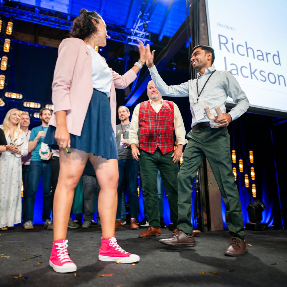 This image shows a joyous moment at a conference where a woman in a blue blazer and pink sneakers is giving a high-five to a man in a green shirt and brown shoes. They are on stage under lights, with an audience applauding in the background. The man's name, Richard Jackson, appears on a screen above him. The scene captures a celebration or award ceremony with a lively and cheerful atmosphere.