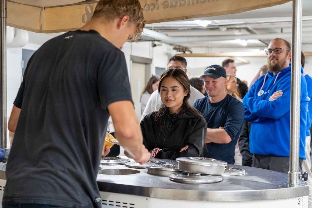 A young woman in a black jacket stands at an ice cream cart, watching as the server scoops ice cream. Behind her, a group of people including a man in a navy hoodie and another in glasses patiently wait their turn.