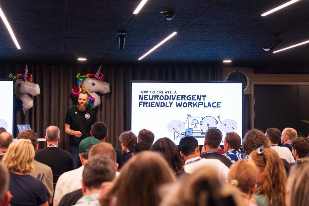 Speaker presenting to a packed audience at an indoor event. The screen behind displays the title 'How to Create a Neurodivergent Friendly Workplace' with cartoon brains. Unicorn balloons decorate the stage.