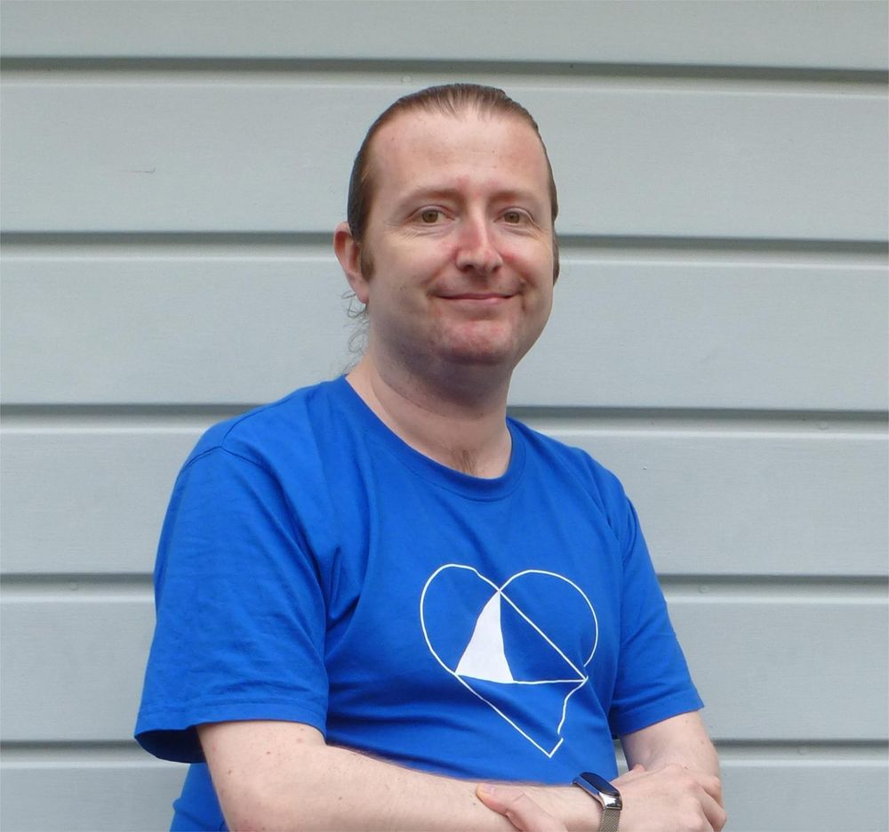 Terence, wearing a blue shirt with a heart-shaped design, smiles while sitting in front of a light gray, paneled background.