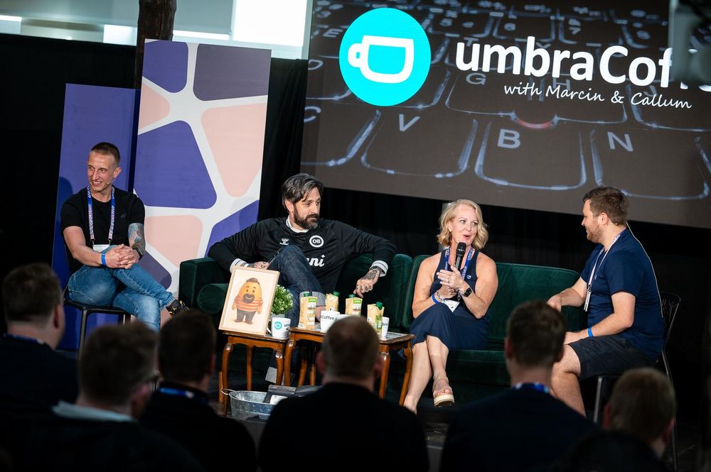 Four speakers on stage during a live recording of 'umbraCoffee with Marcin & Callum' at a Codegarden event. They're seated in front of an audience with branding, coffee drinks, and a plush mascot on display.