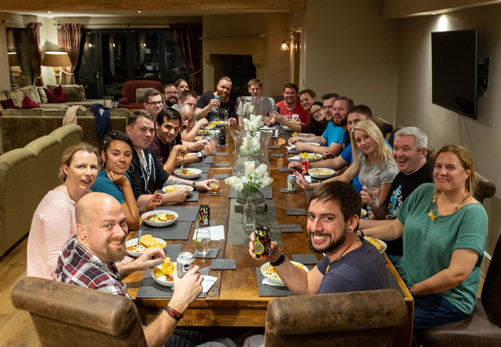 A large group of people sits around a long wooden dining table, smiling and enjoying a shared meal in a cozy indoor setting. Plates of food, drinks, and decorative flowers fill the table, and many attendees raise glasses or cans in a toast. The relaxed atmosphere suggests a retreat or community event.