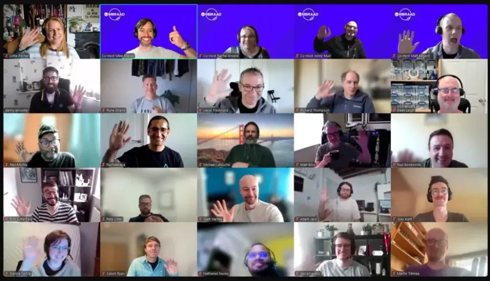 A virtual meeting screenshot featuring a 5x5 grid of smiling participants, all waving at the camera. Each person is framed in their own video call window, with a mix of home office backgrounds and a few virtual ones. Several attendees have “Co-Host” labels and bright purple Umbraco backgrounds, indicating a branded or themed event. The overall mood is warm, friendly, and inclusive.