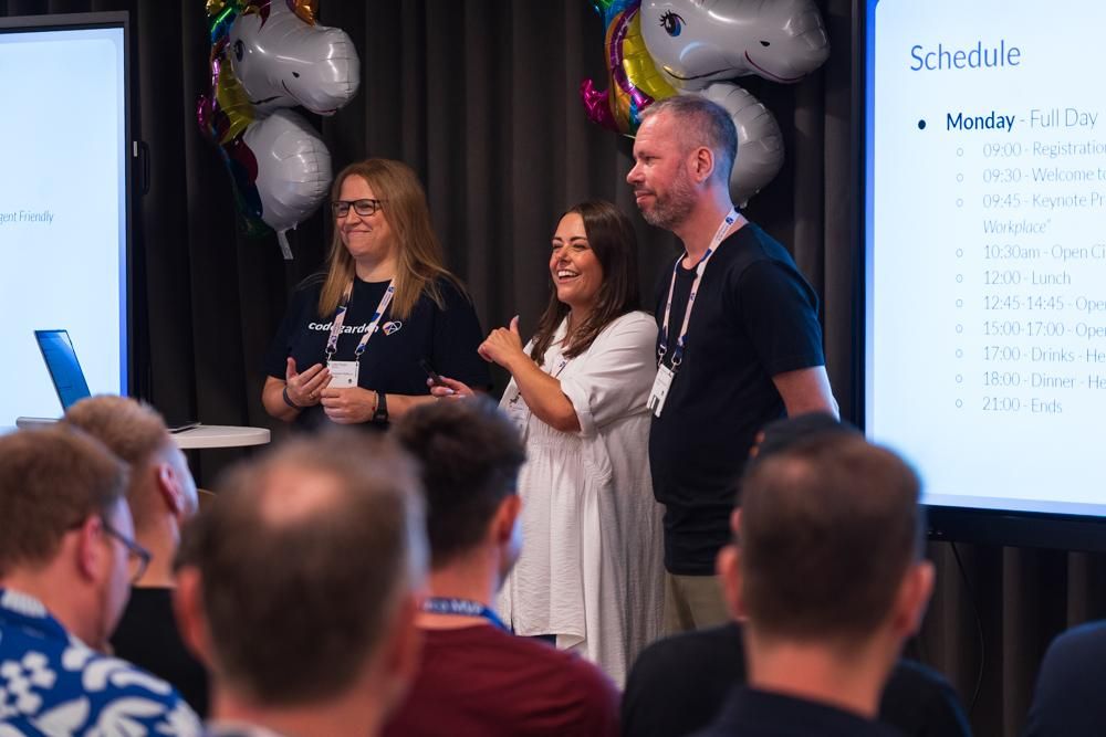 Three speakers standing in front of a projected event schedule, smiling and addressing an audience. The setup includes unicorn balloons and Codegarden branding.