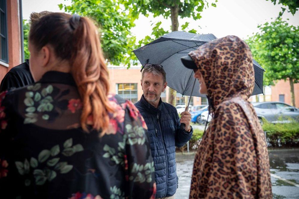 A man holding a black umbrella stands outside talking to two women, one with a floral top and red ponytail, the other in a leopard-print raincoat. It appears to be raining, with wet pavement and water droplets visible on the umbrella and surroundings.