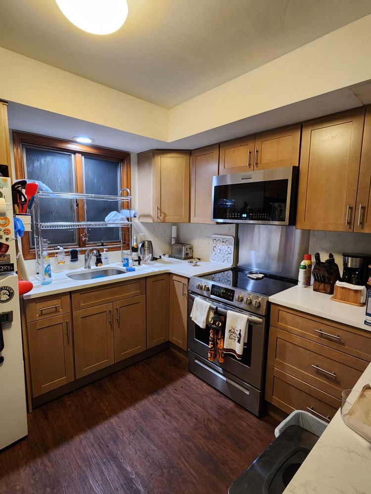 A small kitchen with brown wooden cabinets, white stone countertops and a steel microwave and electric range. 
