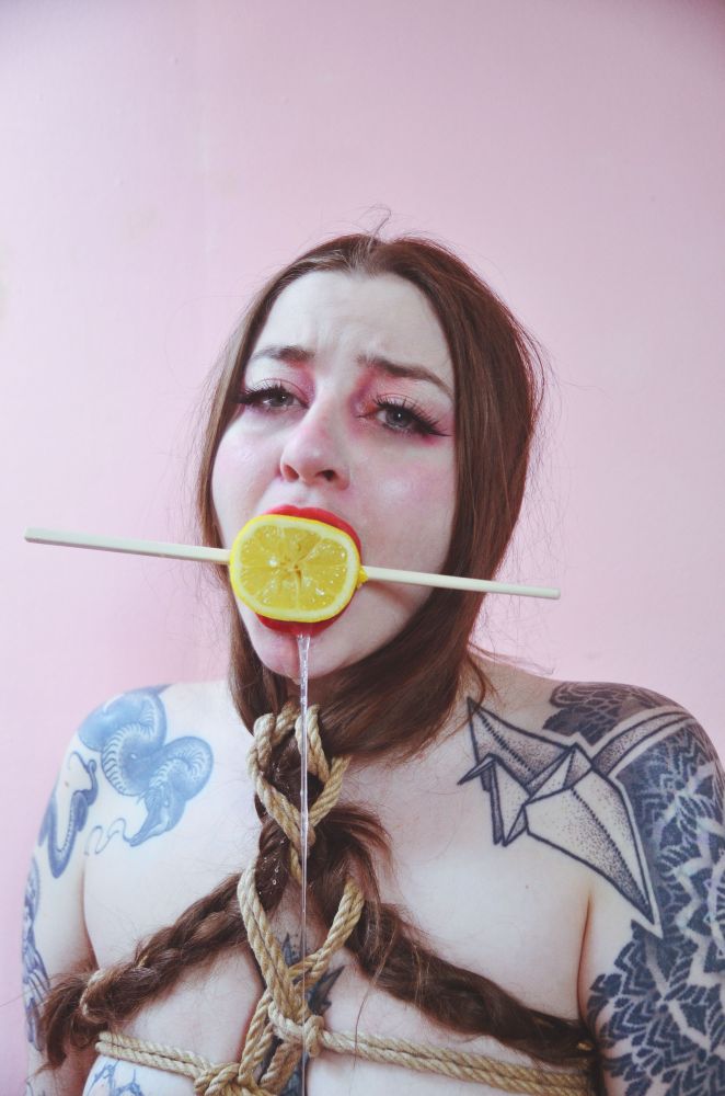 Freshie Juice is bound, her braids put into the ties and she has a lemon in her mouth with a chop stick through it. She is drooling and standing before a pink wall 
