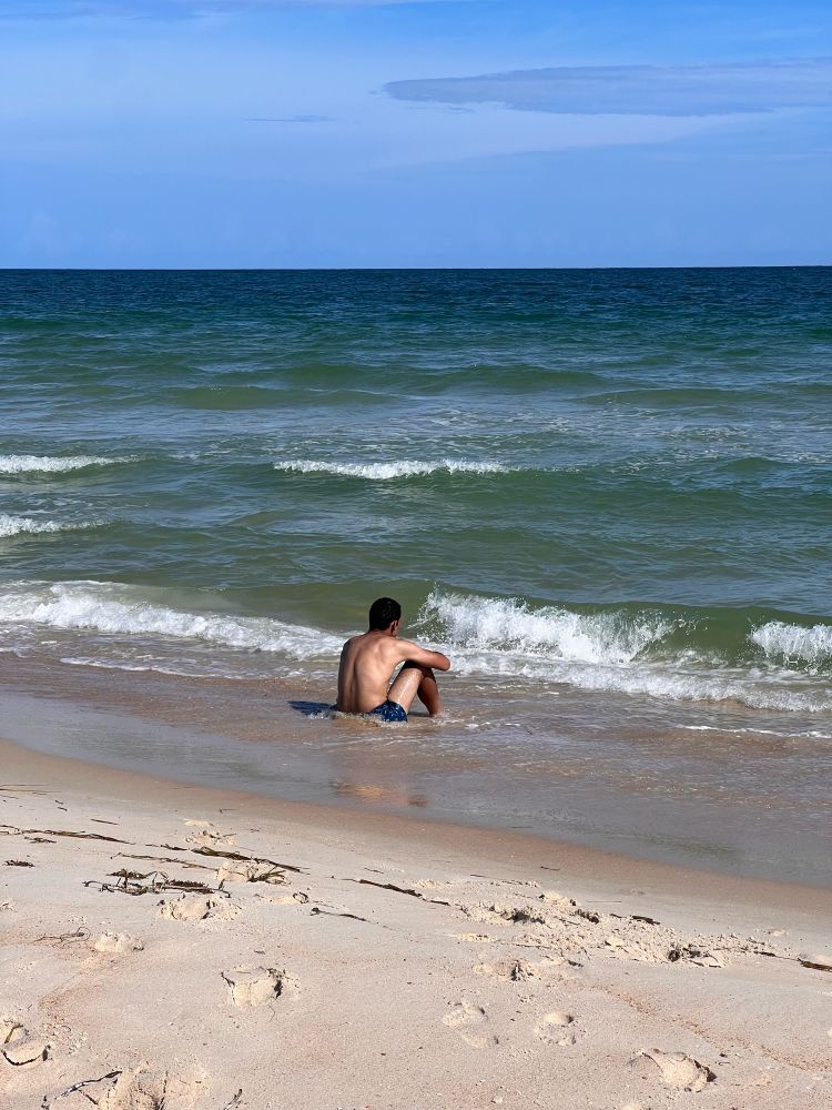 Young man sits on the beach facing away from the camera with soft, blue-green waves rolling in.
