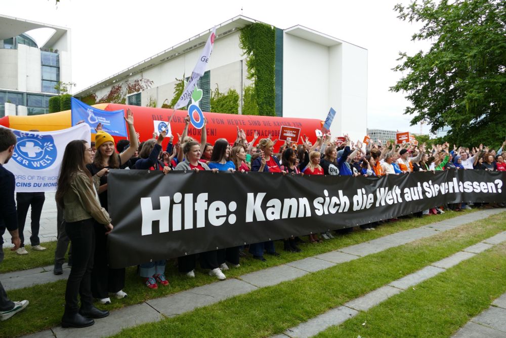 Weitere Perspektive: Eine Menschengruppe demonstriert mit Spruchbändern und Bannern vor dem Bundeskanzleramt in Berlin. Im Hintergrund wird ein riesiger roter Stift hochgehalten, darauf die Aufschrift: "Die Kürzungen von heute sind die Krisen von morgen! #LuftNachOben"
