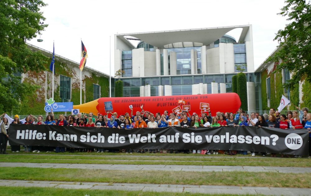 Eine Menschengruppe demonstriert mit Spruchbändern und Bannern vor dem Bundeskanzleramt in Berlin. Im Hintergrund wird ein riesiger roter Stift hochgehalten, darauf die Aufschrift: "Die Kürzungen von heute sind die Krisen von morgen! #LuftNachOben"
