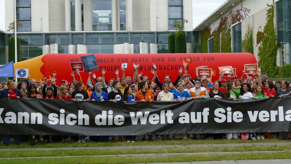 Andere Perspektive: Eine Menschengruppe demonstriert mit Spruchbändern und Bannern vor dem Bundeskanzleramt in Berlin. Im Hintergrund wird ein riesiger roter Stift hochgehalten, darauf die Aufschrift: "Die Kürzungen von heute sind die Krisen von morgen! #LuftNachOben"
