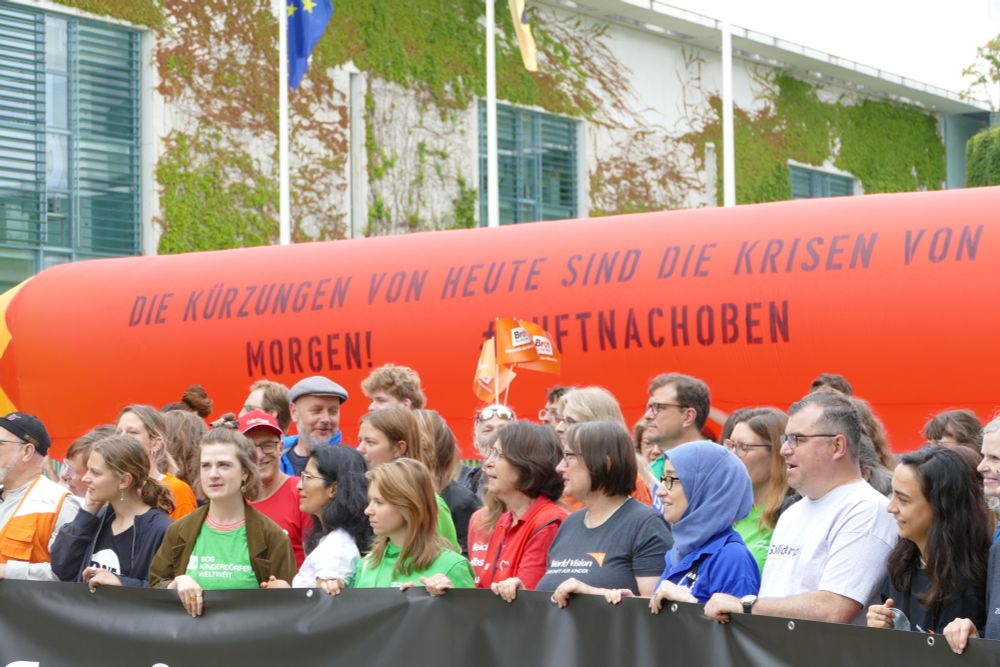 Andere Perspektive: Eine Menschengruppe demonstriert mit Spruchbändern und Bannern vor dem Bundeskanzleramt in Berlin. Im Hintergrund wird ein riesiger roter Stift hochgehalten, darauf die Aufschrift: "Die Kürzungen von heute sind die Krisen von morgen! #LuftNachOben"
