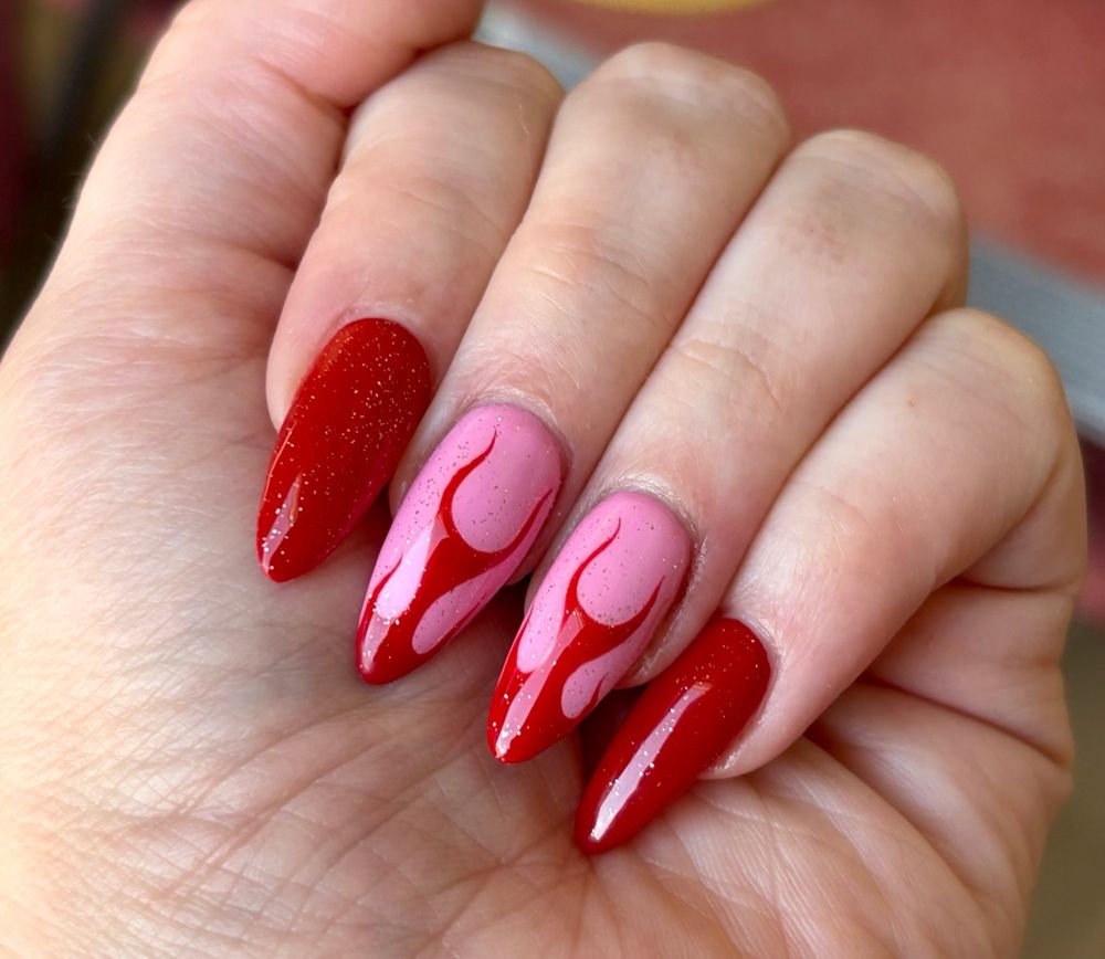 A hand clenched into a fist to show off a set of long red nails with the two middle fingers featuring a pink background and red flames 