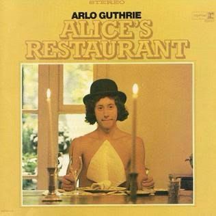 The cover of Arlo Guthrie’s Alice’s Restaurant album. Arlo is seated at a table and he’s shirtless, with a napkin on his chest. There are two candles on the table. His name and the title are above the photo.