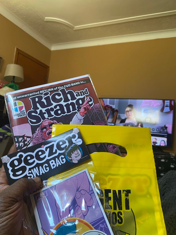 Allan is holding the contents of his Geezer comic Kickstarter fulfillment. Pictured is a Geezer Swag Bag, issue 3 of Geezer in a yellow Argent Studios bag, and the added-on Rich and Strange comic. A video from harpist Emily Hopkins is playing in the background.