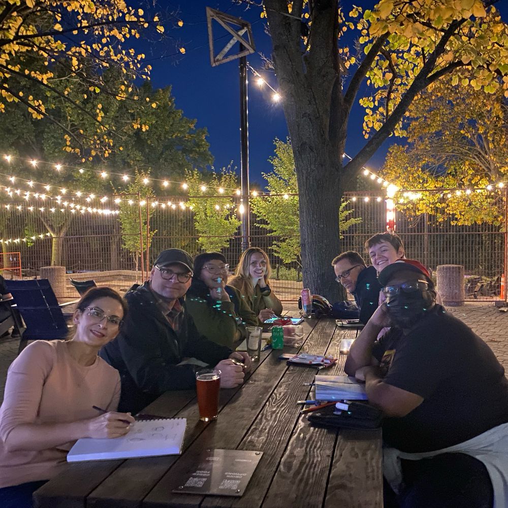 The DrawMob gang, outside at The Forks. The group of seven people of various ethnicities are at a table and lit by outdoor lights.