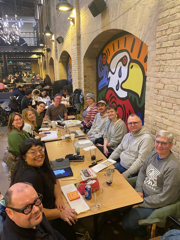 Eleven people (of multiple ethnicities) of the DrawMob collective sitting at a table at The Forks Market in Winnipeg.