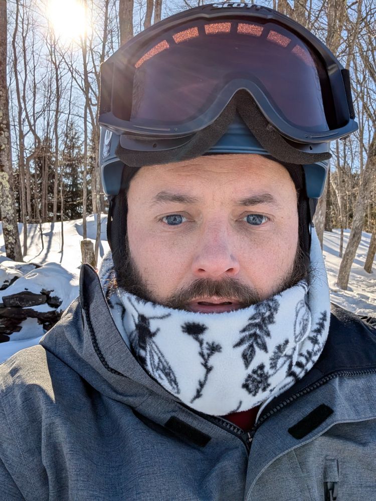 Selfie of the poster in a snowboarding helmet and black and white neck warmer with trees behind him. 